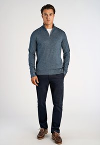Marineblaues Zip-Pullover mit strukturiertem Finish, getragen über einem weißen Hemd, kombiniert mit dunklen Hosen und braunen Bootsschuhen.