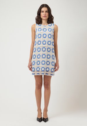 Vrouw staand, gekleed in een mouwloze gehaakte jurk met blauwe en beige bloemenpatronen en zwarte schoenen met spitse neus op een effen achtergrond.