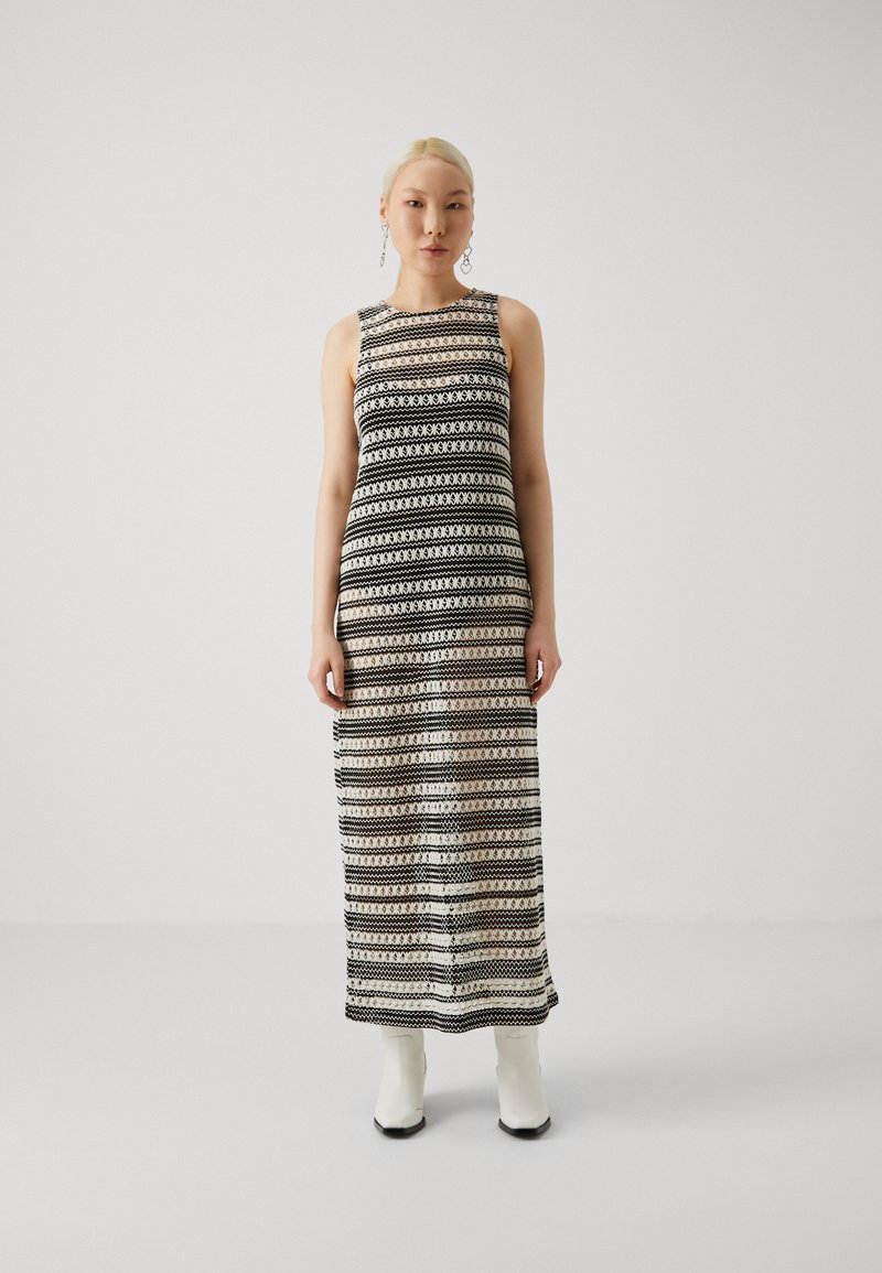 Robe maxi sans manches présentant un motif géométrique noir et blanc, avec une silhouette fluide et un design fendu, associée à des bottes blanches aux chevilles.