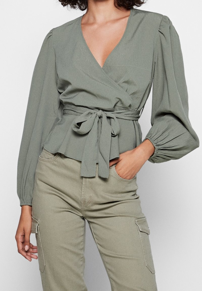 Femme portant un chemisier cache-cœur vert sauge avec des manches longues bouffantes et une taille nouée, associé à un pantalon cargo vert clair.