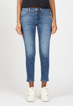 Schlanke blaue Jeans aus Denim mit normaler Taillenhöhe, ausgestattet mit fünf Taschen und einem gekürzten Saum am Knöchel mit leicht ausgefransten Kanten. Kombiniert mit weißen Sneakern.