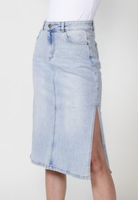 Svetlomodra denim krila z oprijetenim pasom, dolžine do kolen in stranskim razporkom. Ima pet žepov in klasično zaklepanje na gumb.