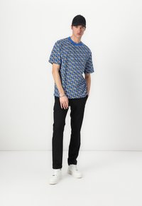 Kurzärmliges blaues Hemd mit einem weißen und schwarzen geometrischen Muster, kombiniert mit schwarzen Hosen und weißen Sneakers, abgerundet mit einer schwarzen Kappe.