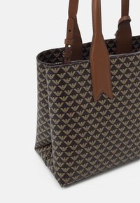 Hnědá taška přes rameno s vzorem a zlatým emblémem, doplněná hnědými koženými detaily a nastavitelnými popruhy. Strukturovaný tvar a pevná textura.