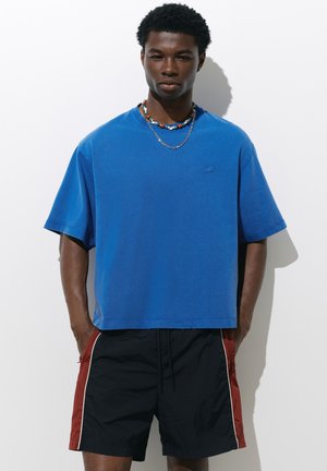 Jeune homme aux cheveux courts et bouclés portant un t-shirt bleu oversize, un short noir avec des côtés rouges et deux colliers sur fond blanc.