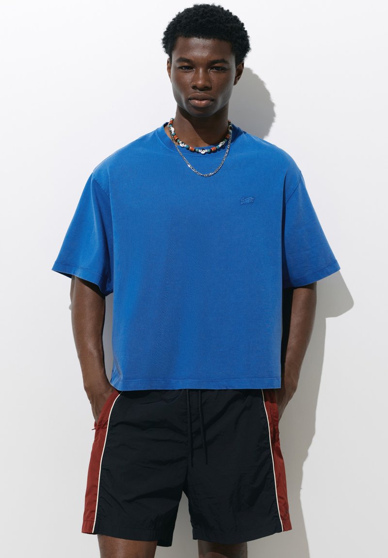Jeune homme aux cheveux courts et bouclés portant un t-shirt bleu oversize, un short noir avec des côtés rouges et deux colliers sur fond blanc.