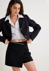 Musta lyhythihainen blazer, jossa on suuret napit, päällä valkoinen raitapaita. Mustat korkeavyötäröiset shortsit, joissa on kontrastina valkoinen reunus.