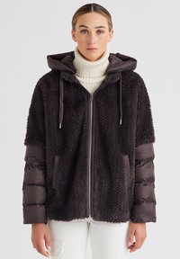 Dunkelviolette Daunenjacke mit einem flauschigen, strukturierten Körper, glatten Ärmeln, Kapuze, Reißverschluss und Fronttaschen. Getragen über einem weißen Rollkragenpullover.