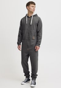Šedý kombinéz s kapucňou na zips, vyrobený z mäkkej látky; má predné vrecko a malú béžovú nášivku s logom; nosí sa s čiernymi teniskami.