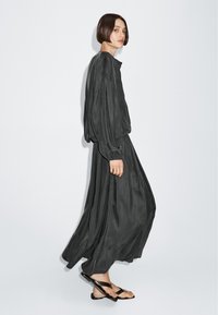 Vrouw met kort haar die een losse donkergrijze blouse met lange mouwen draagt, bijpassende wijde rok en zwarte sandalen, tegen een effen witte achtergrond.