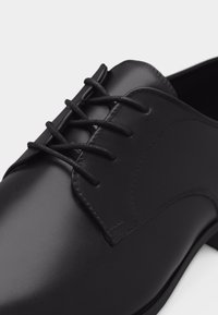 Zapato de vestir de cuero negro con cierre cerrado y acabado liso, mostrando costuras detalladas y punta redondeada.