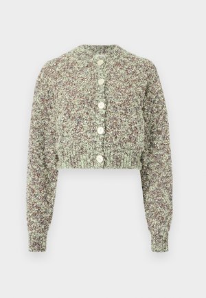 Femme portant un cardigan beige texturé, un top marron, un pantalon ample vert clair, des talons blancs, tenant un grand sac à main marron foncé, debout sur un sol gris.