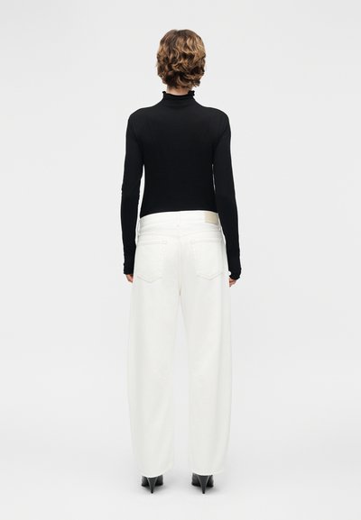 Άτομο με κοντά, σγουρά μαλλιά που φοράει μαύρο μακρυμάνικο τοπ, λευκό παντελόνι με φαρδιά μπατζάκια και μαύρα παπούτσια με μυτερή φτέρνα, στέκεται γυρισμένο προς τα πίσω.