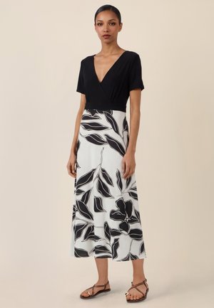 Frau steht, trägt ein schwarzes, kurzärmeliges V-Ausschnitt-Oberteil und einen weißen Rock mit großen schwarzen Blumenmustern, dazu braune Riemchensandalen.