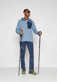 Hele sinine lukuga jakk tumesinise rindtaskuga, koos tumesiniste pükstega. Kandmas päikeseprille, hoides matkakeppe ning sobib välitegevusteks.