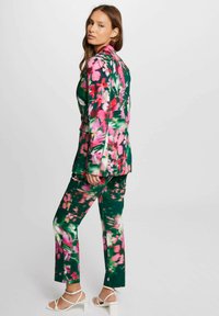 Completo floreale con base verde e fiori rosa e bianchi. La giacca ha un colletto a punte e i pantaloni sono leggermente svasati con una texture liscia.