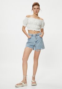Biała bluzka typu crop top bez ramiączek z falbanistymi krawędziami, w połączeniu z jasnoniebieskimi szortami dżinsowymi z postrzępionymi brzegami i solidnymi kieszeniami. Beżowe sandały na platformie.
