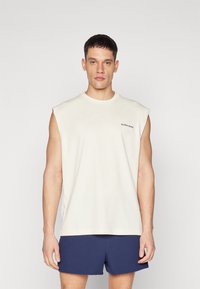Light beige sleeveless t-shirt with a rounded neck, made of soft cotton. Features a small black logo on the chest.