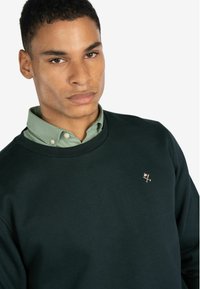 Sudadera verde oscuro con cuello redondo y un pequeño logo bordado en rosa claro, superpuesta a una camisa de color verde claro con cuello.