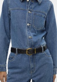 Macacão de ganga em azul claro, com fecho frontal de botões e bolso no peito; combinado com um cinto de pele preto com uma fivela em tom dourado.