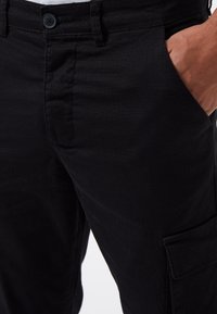 Pantalones cargo negros con una tela texturizada, que presentan un cierre de botón, bolsillos laterales y detalles de costura visibles.