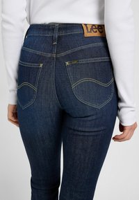 Mörkblå denimjeans med hög midja, gula sömmar och en brun läderlogotyp på baksidan av midjebandet.