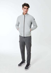 Giacca zip-up grigia con alto collo, vestibilità slim. Abbinata a pantaloni cargo grigio scuro e sneakers bianco e nere. Tessuto liscio, senza pattern.