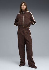 Γυναίκα στέκεται με τα χέρια στις τσέπες, φορώντας σκούρο καφέ φόρμα Puma με κρεμ ρίγες στα μανίκια και το παντελ όνι, μπροστά σε γκρίζο φόντο.