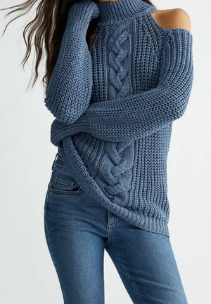 Mujer con cabello largo, vistiendo un suéter azul de punto con hombros descubiertos y jeans azules, posando con un brazo cruzado sobre la cintura y la mano cerca del rostro.