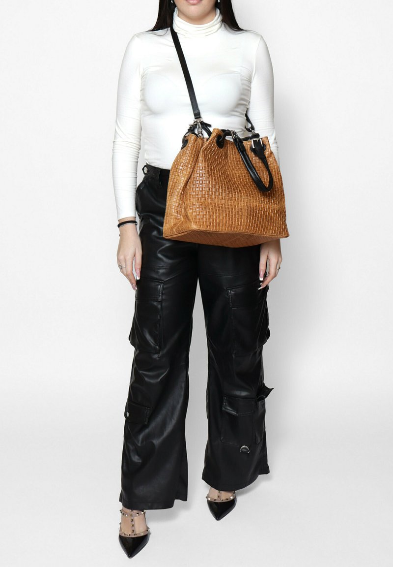 Huisgemaakte, geweven tas in een tan kleur met zwarte leren banden, met een gestructureerd oppervlak. Model draagt zwarte leren cargobroek en een wit shirt met lange mouwen.