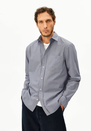 Homme aux cheveux courts et bouclés portant une chemise à carreaux bleue boutonnée par-dessus un t-shirt blanc, debout les mains dans les poches.