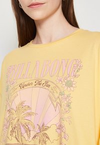 Camiseta de algodón amarilla con un gran gráfico rosa que presenta diseños florales y palmeras. Texto "BILLABONG" encima de "Bajo el sol."