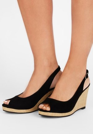 Pies con sandalias cuña de tiras negras y punta abierta con plataforma tejida y texturizada sobre fondo blanco.