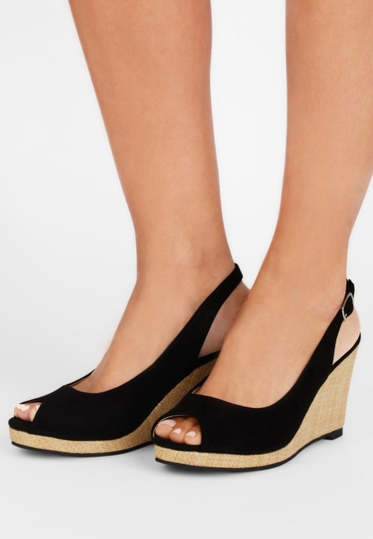 Pieds portant des sandales compensées à bride arrière noires ouvertes à l'avant avec des talons plateforme tressés texturés sur fond blanc.