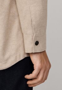 Camisa beige con un tejido texturizado, cierre de botón visible en el puño y un dobladillo bien cosido. Pantalones oscuros parcialmente visibles.