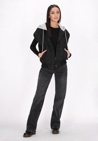 Gilet trapuntato nero con cappuccio grigio, chiusura con zip e due tasche frontali. Abbinato a jeans a gamba larga grigio scuro e scarpe nere.