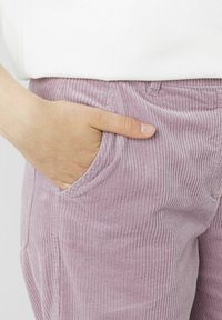 Pantalons en velours côtelé de couleur lavande claire avec des nervures verticales, comportant des poches avant et une coupe décontractée. Tissu texturé montré près d'une main.