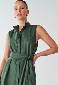 Roheline puuvillane kleit seistekaelusega, nööbiga esiosal, lühikeste varrukatega ja seotud vööga. Omab rinna-taskut ja plisseeritud seelikut.