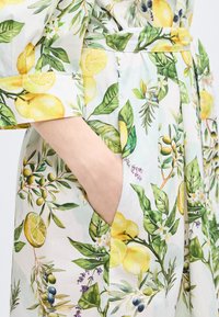 Main dans la poche d'une robe blanche ornée d'un imprimé de citrons jaunes et de branches d'olivier vertes avec feuilles et petites fleurs.