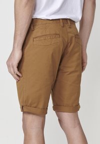 Man som bär bruna shorts med upprullade fållar och en vit t-shirt, visad bakifrån, stående mot en enkel bakgrund.