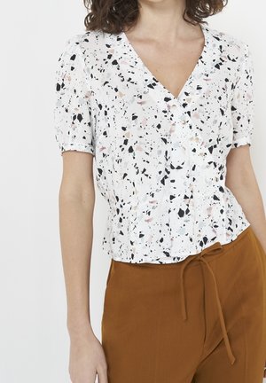 Woman wearing white short-sleeve blouse with black and pink speckles and brown high-waist pants with a drawstring waist.