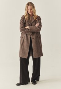 Abrigo de lana marrón con diseño de espiga, de doble botonadura, grandes bolsillos y cierre de dos botones, combinado con pantalones negros holgados.