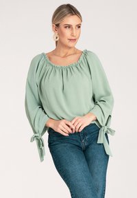 Jasnozielona bluzka z okrągłym dekoltem, bufkowymi rękawami i wiązanymi akcentami. Zestawiona z ciemnoniebieskimi dżinsami o gładkiej fakturze.