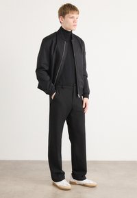 Black bomber jacket with a smooth texture, worn over a black turtleneck, paired with black trousers and white sneakers. Minimalist design.
