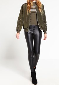 Olivově zelená bomber bunda přes tmavě pruhovaný svetřík, v kombinaci s černými koženými legínami a kotníkovými botami, ukazuje elegantní a obepnutou siluetu.