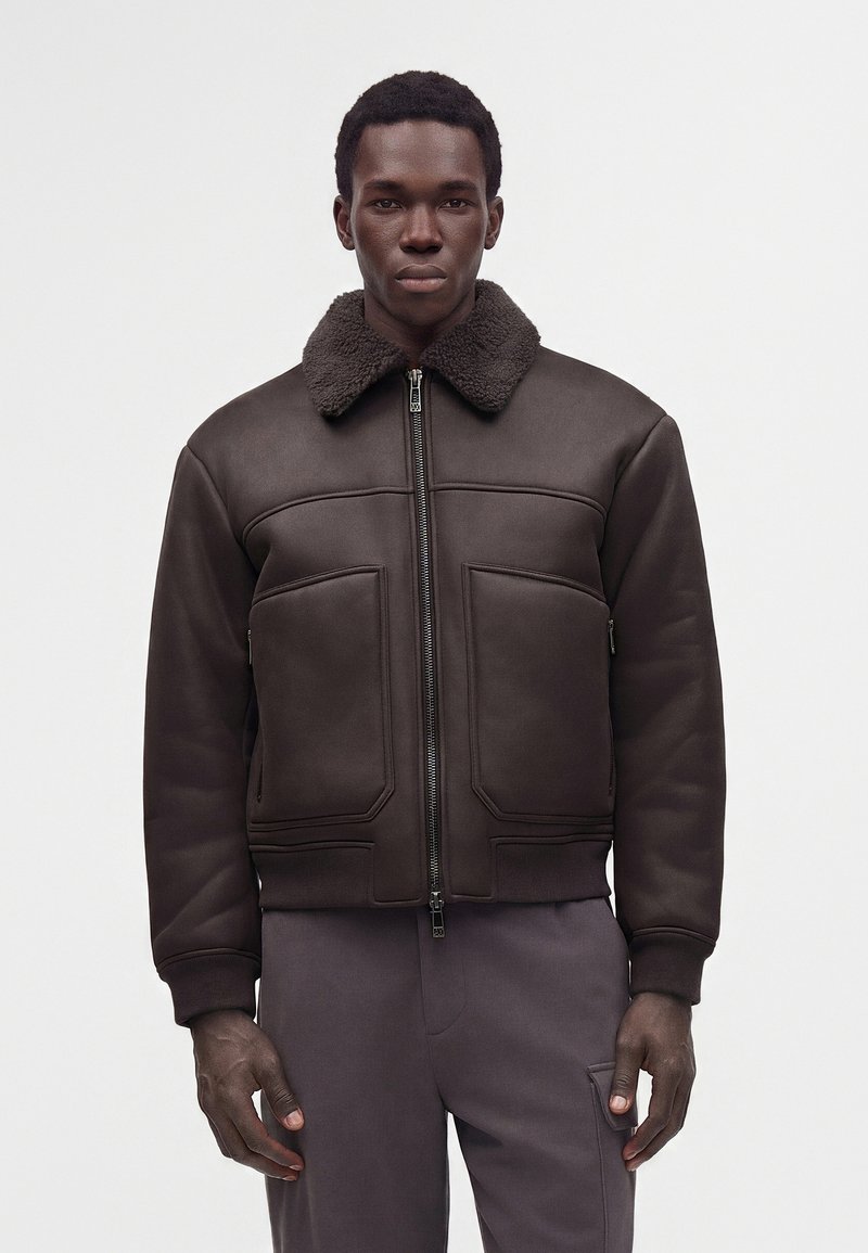Homme portant une veste zippée marron foncé avec un col en peau lainée et de grandes poches avant, associée à un pantalon gris foncé sur un fond uni.