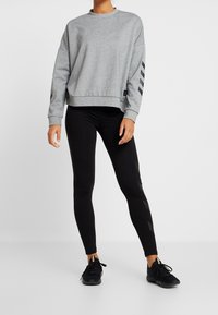 Felpa grigia con maniche lunghe e strisce nere sulle braccia, abbinata a leggings neri e scarpe da ginnastica nere.