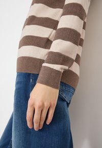 Main reposant sur le côté, portant un pull rayé marron et beige ainsi qu'un jean en denim bleu, sur un fond uni.
