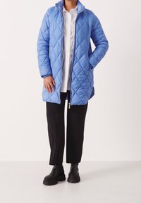 Cappotto lungo trapuntato di un blu chiaro con colletto alto, chiusura con zip e due tasche laterali, abbinato a pantaloni neri e stivali neri robusti.