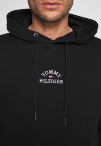 Black hoodie featuring a drawstring with silver tips, embroidered "TOMMY HILFIGER" logo in white and red, made from soft fabric.